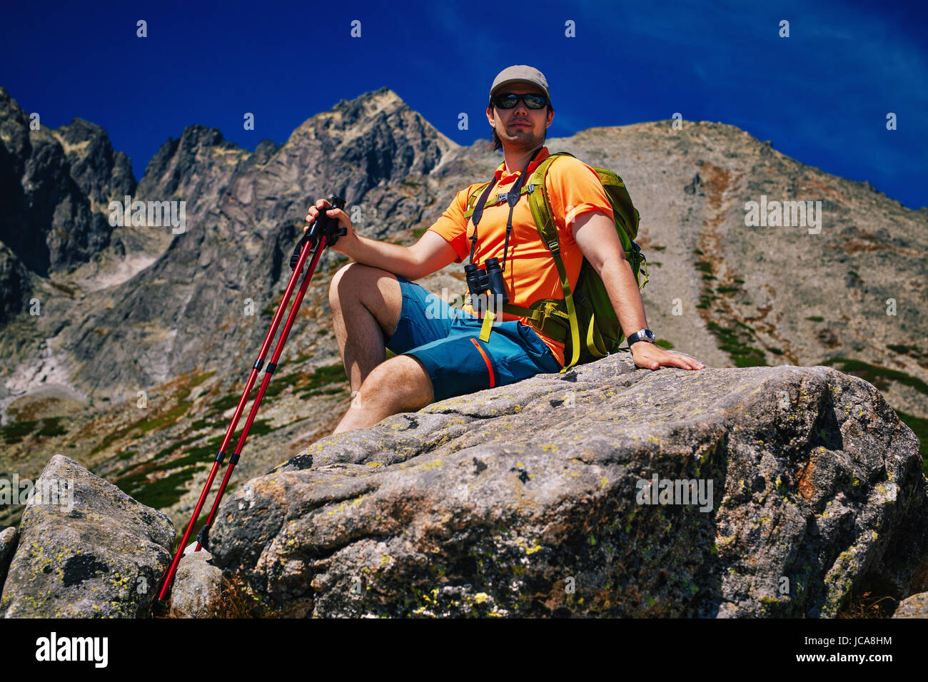 Young man tourist with green backpack and poles sitting at high mountains Stock Photo