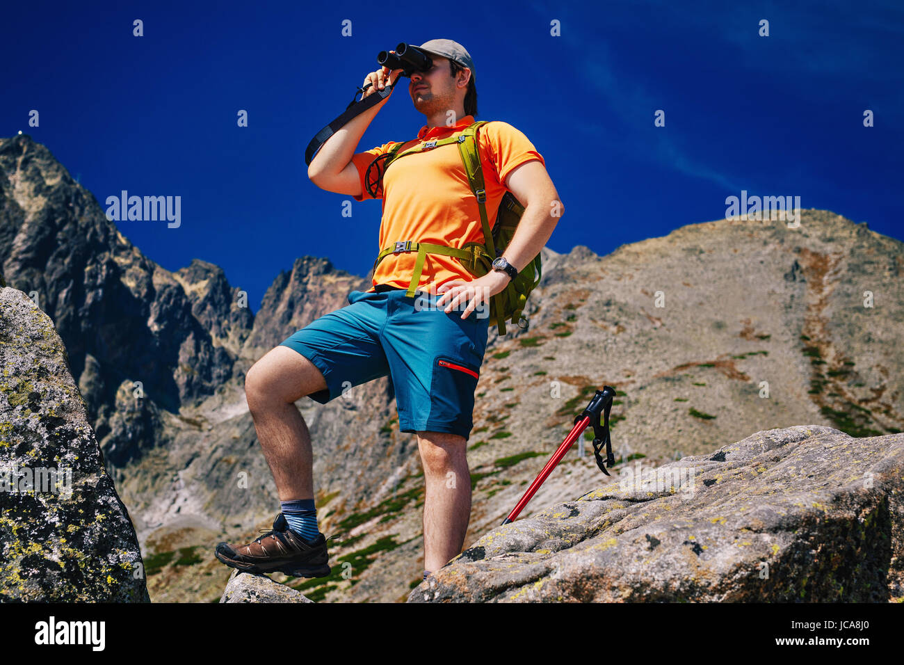 Young man tourist with green backpack looking in binoculars on high mountains Stock Photo
