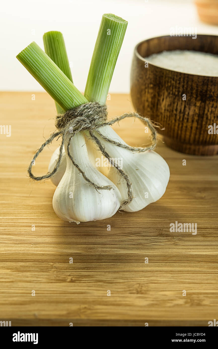 Three garlic bulbs tied up together with packthread on cutting board near the bowl full of salt. Unfocused background. Stock Photo