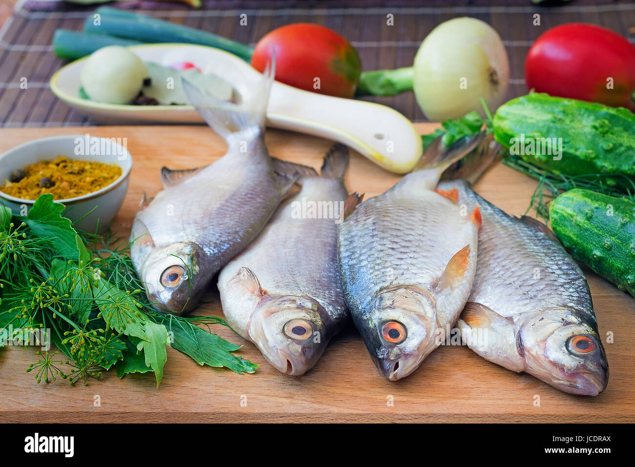 On a table on a chopping board there are a fish, cucumbers, tomatoes, onions, spices and parsley greens. Stock Photo
