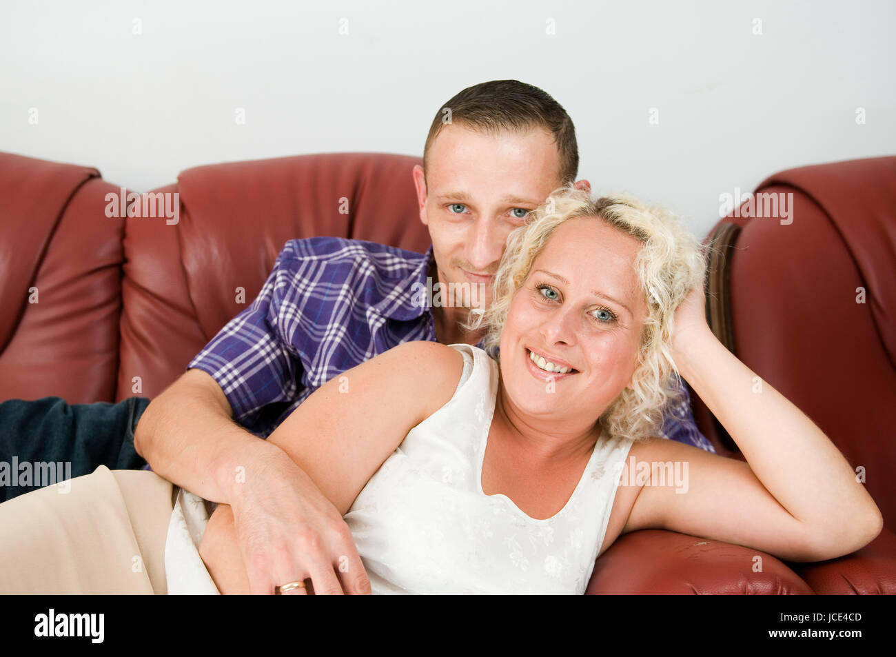 Oberkörper-Porträt eines jungen Paares hintereinander auf einem braunen Leder-Sofa liegend und glücklich in die Kamera lächelnd Stock Photo
