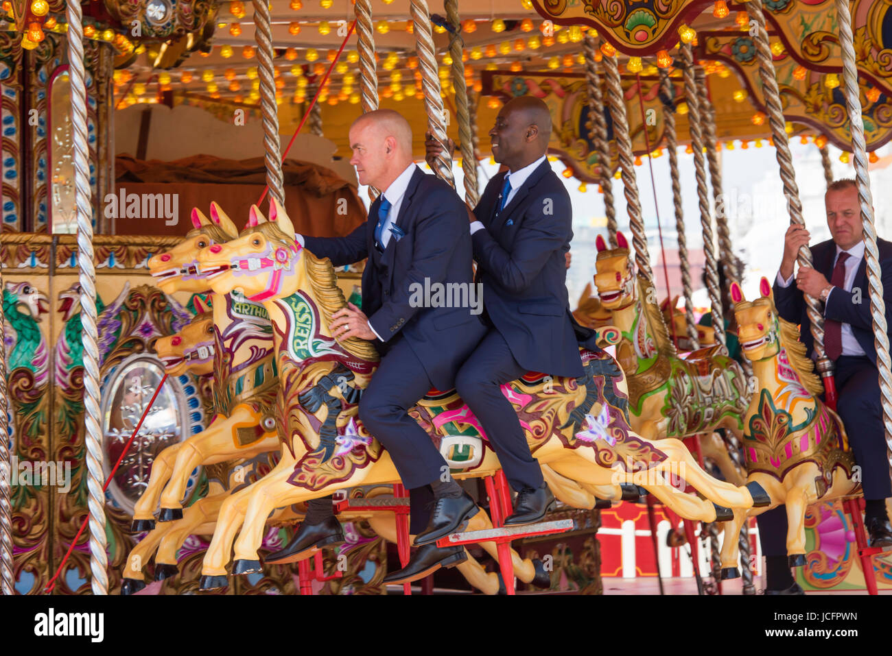 Group of friends wearing suits and dresses on a merry go round on the seafront at Brighton on a sunny day. It looks as if they are part of a wedding Stock Photo