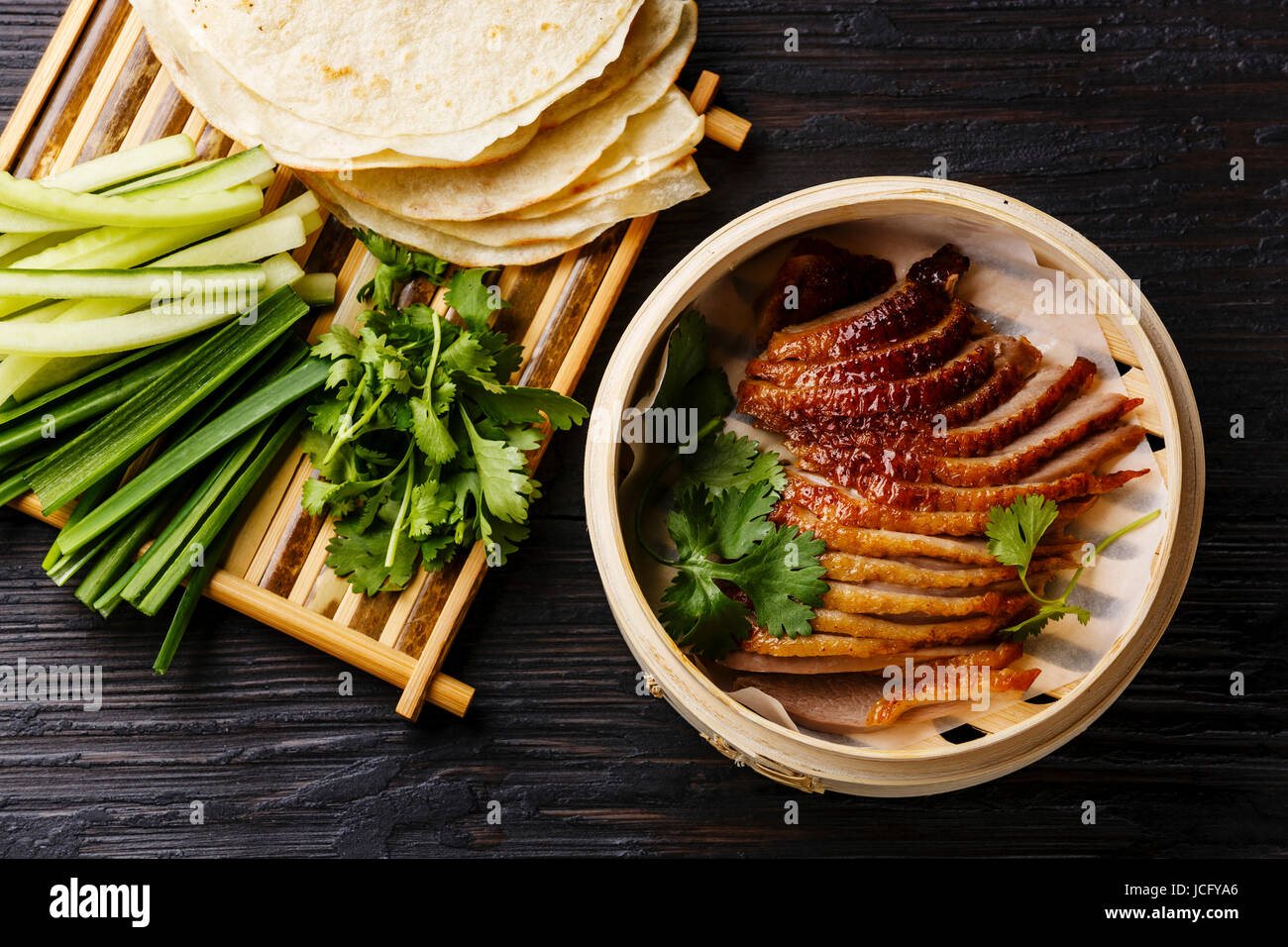 Peking Duck in bamboo steamer served with fresh cucumber, green onions, cilantro and roasted wheaten chinese pancakes on black burned wooden backgroun Stock Photo