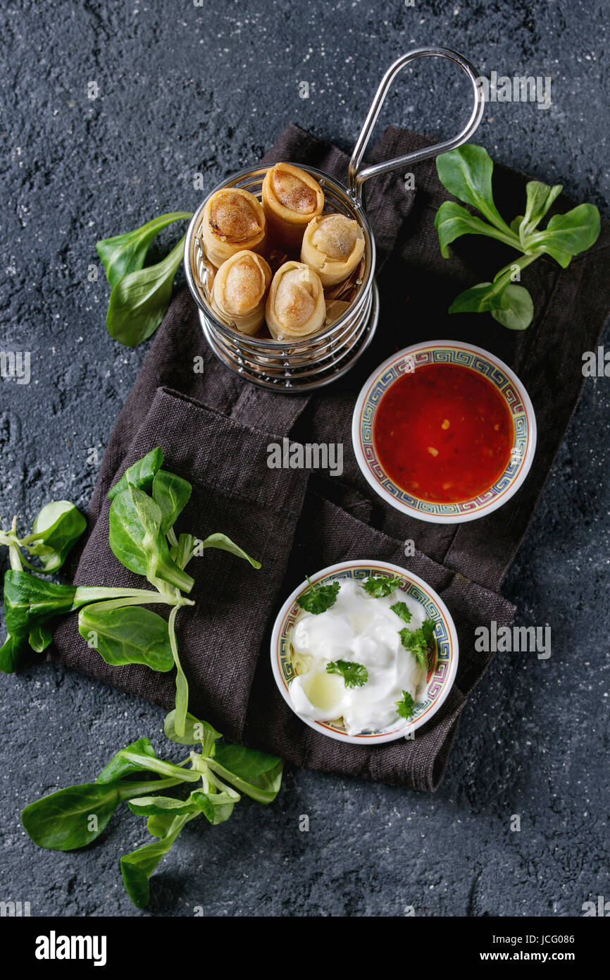 Fried spring rolls with red and white sauces, served in traditional china plate and fries basket with fresh green salad over black texture background. Stock Photo