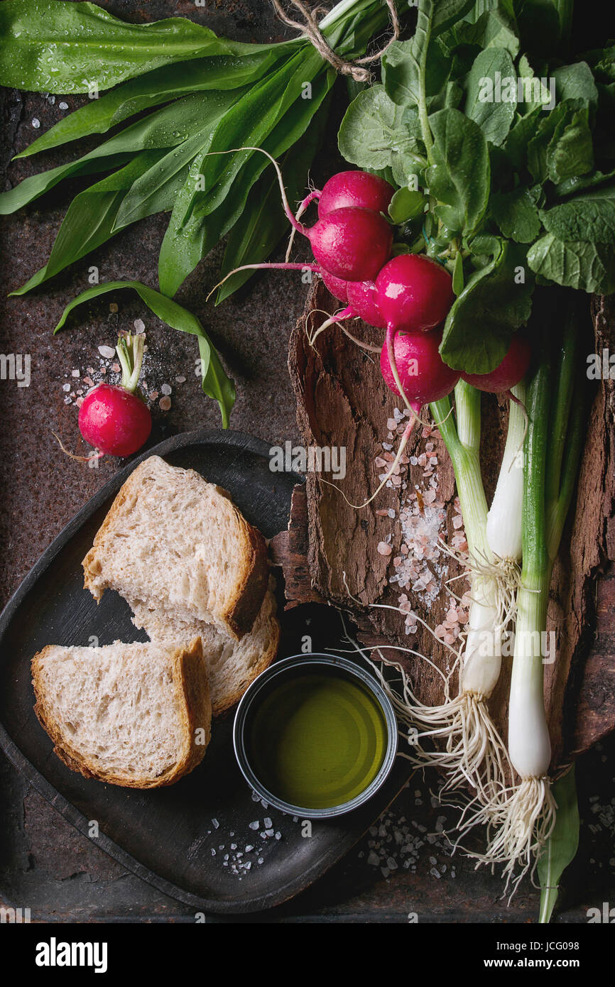 Rustic lunch breakfast with fresh young vegetables radish, spring onion, garlic leaves, salt, olive oil and bread on wooden bark over dark texture met Stock Photo