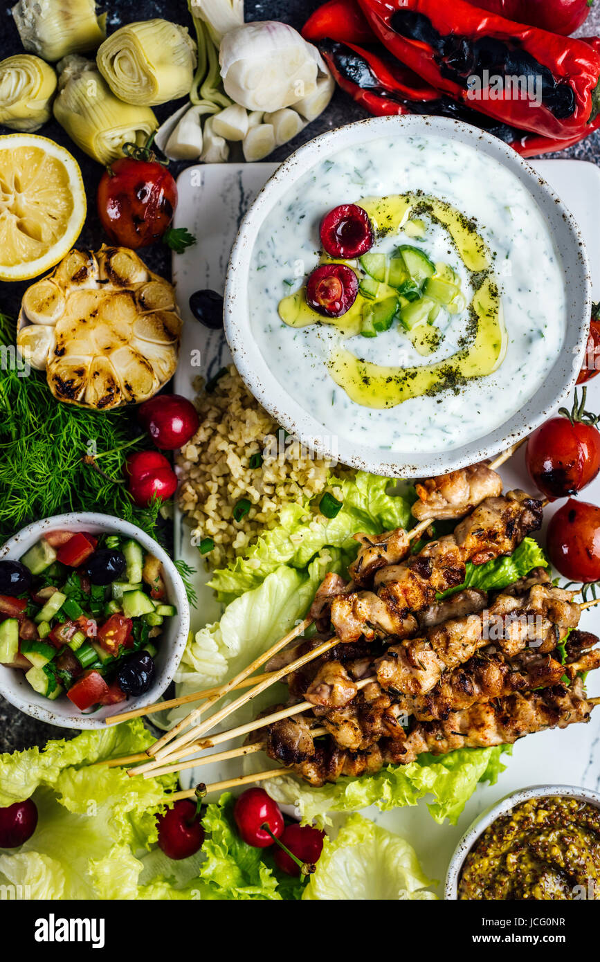 Chicken skewers on a white board served iwth a bowl of cacik, a yogurt and cucumber dip and with various snacks photographed from top view. Stock Photo