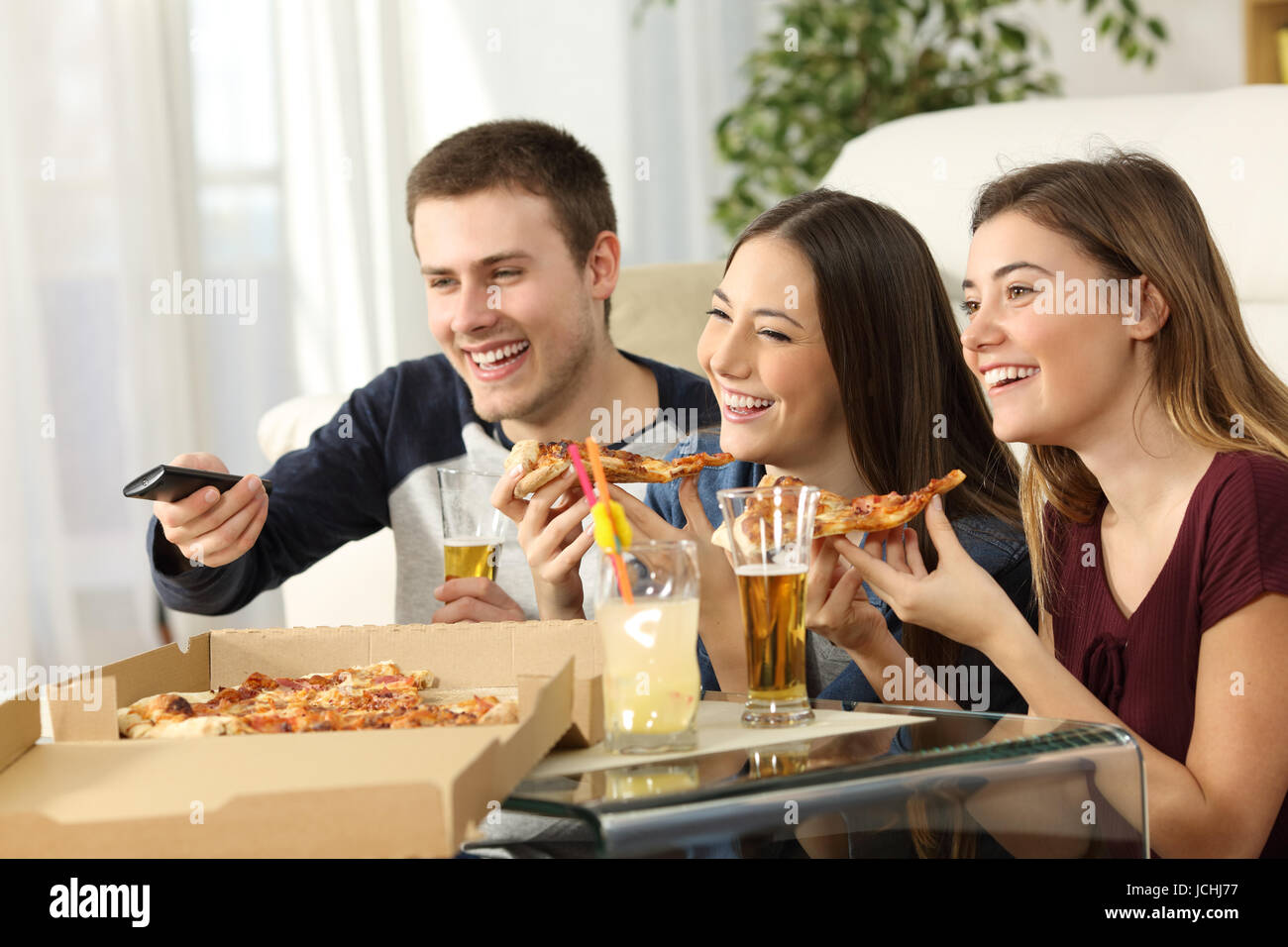 Three friends watching tv and eating pizza sitting on the floor in the living room in a house interior Stock Photo