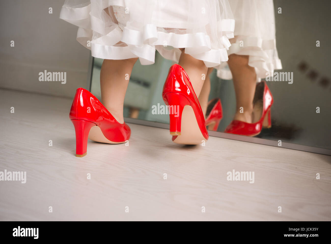 Cute funny little baby girl walks at home trying mom's red high heel shoes Stock Photo