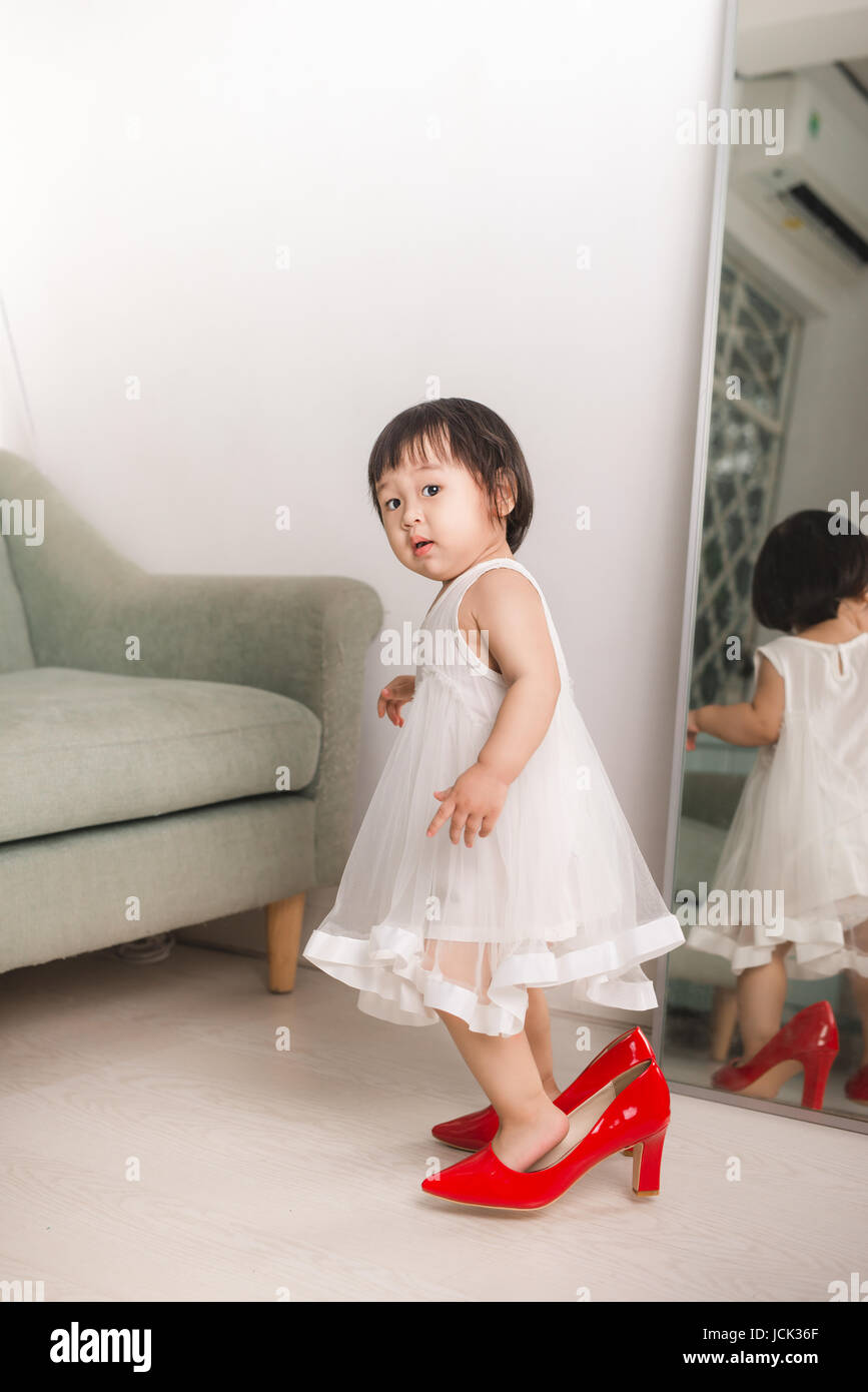 Cute funny little baby girl walks at home trying mom's red high heel shoes Stock Photo