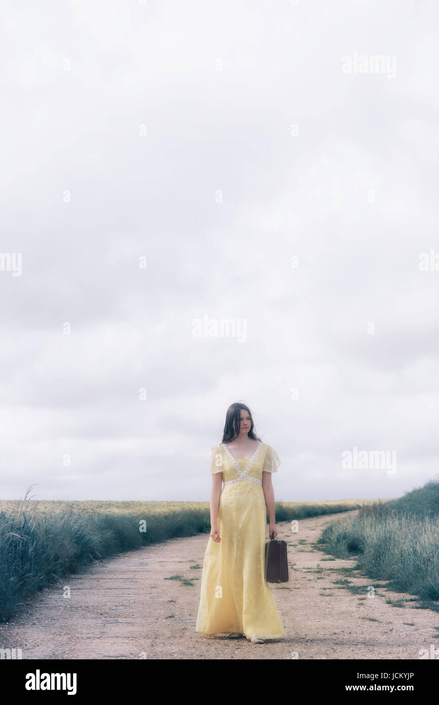 a young girl with a yellow dress is carrying a suitcase and is standing on a path through grainfields Stock Photo