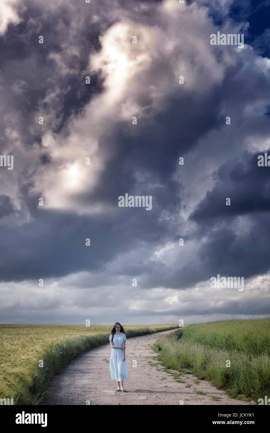 a woman in a blue dress is walking on a path through grainfields under heavy storm clouds Stock Photo
