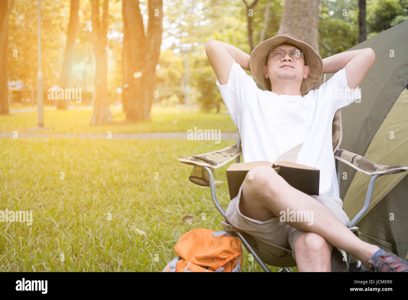 young man tourist sitting on chair resting and relaxing in front of tent at camping site in forest. Outdoor activity in summer. Adventure traveling in Stock Photo