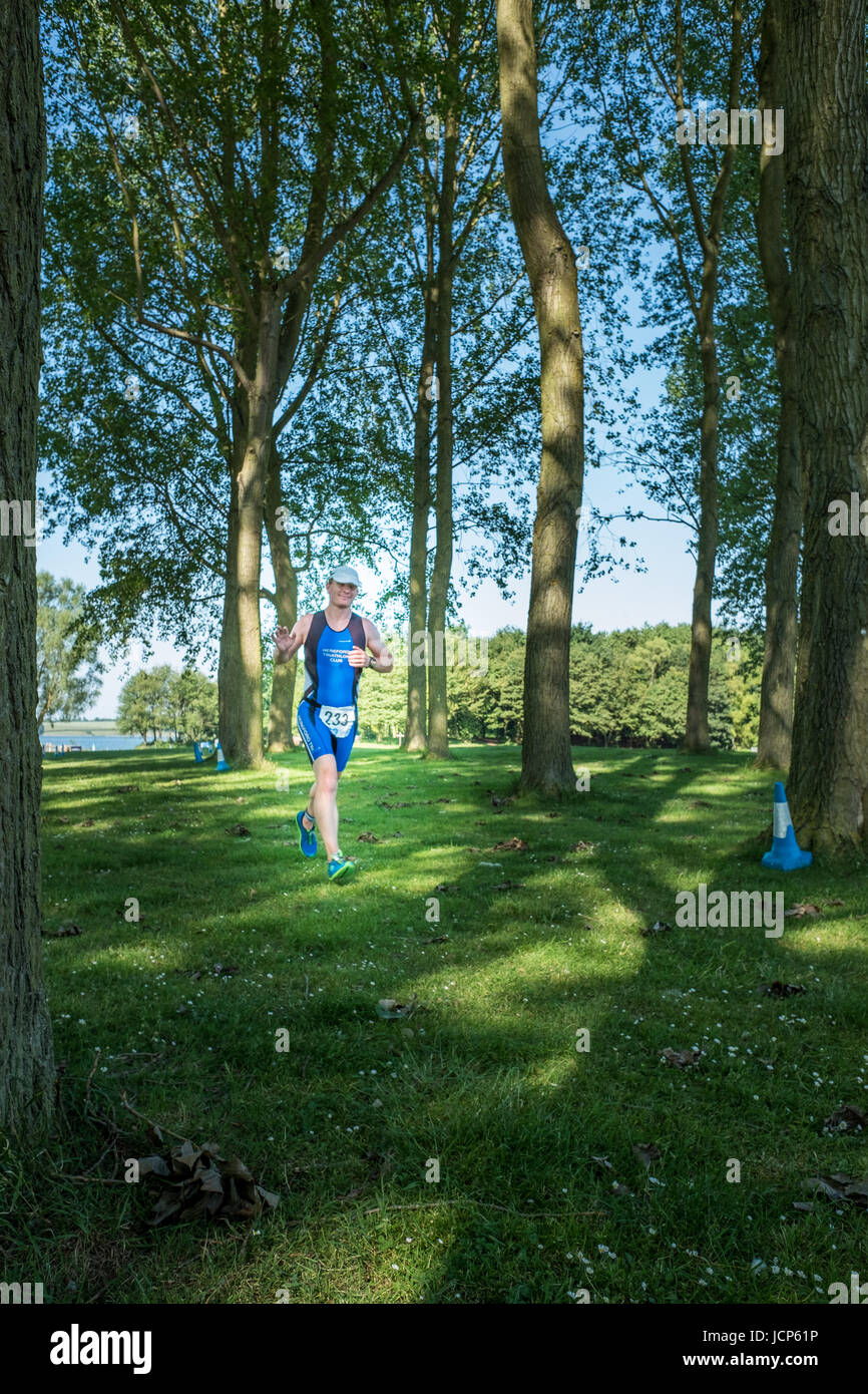 Rutland Water, UK. 17th June, 2017. Iain Darwood, a cheerful, relaxed and tired competitor, gives a wave as he trots through a woodland along the shore of Rutland reservoir on a hot, sunny day during the10 km run, the third and final phase of the Dambuster Triathlon (swim, bike and run race) at Rutland Water, England, on 17 June 2017. Credit: Michael Foley/Alamy Live News Stock Photo