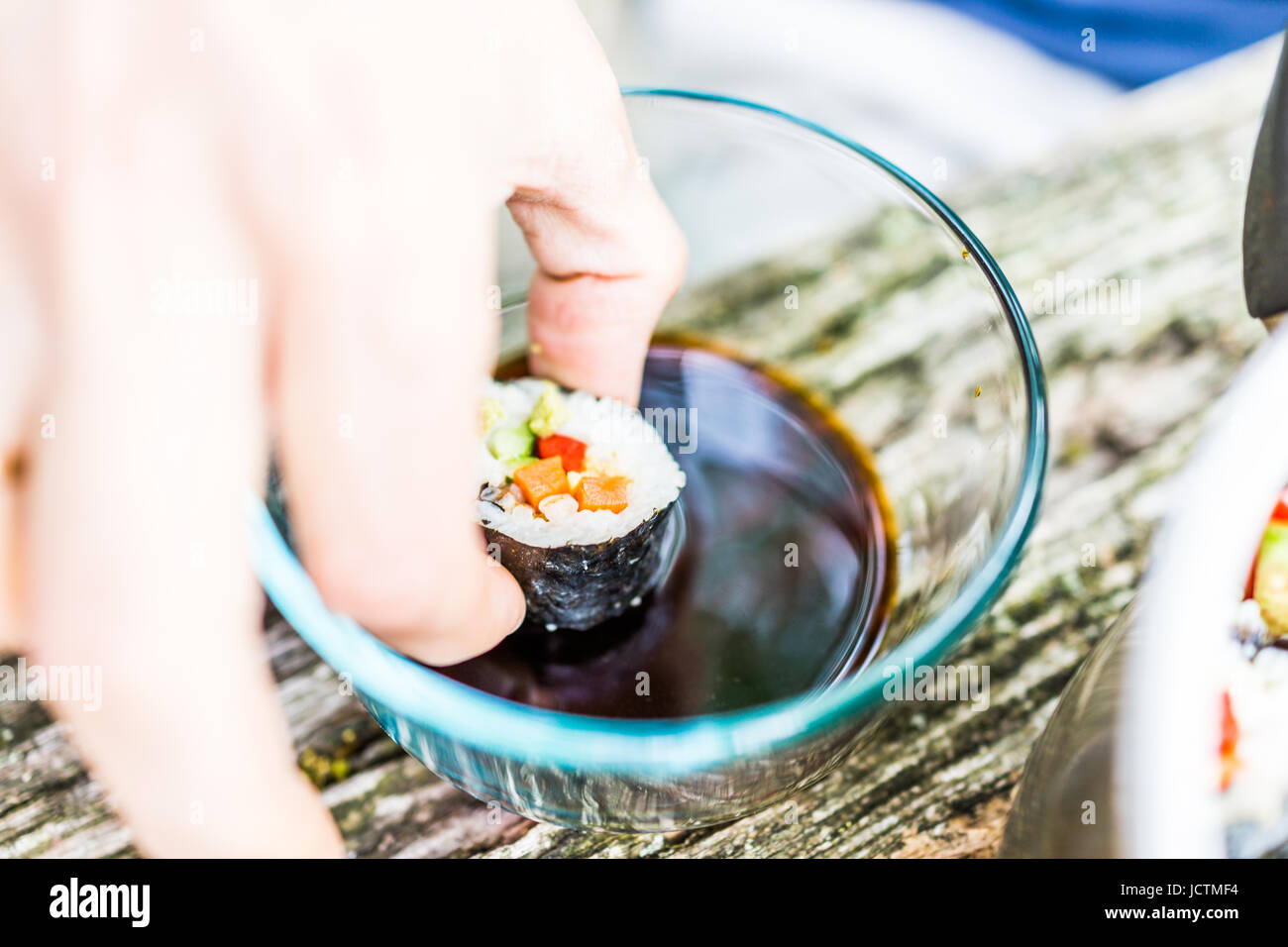 Macro closeup of hand dipping maki sushi in soy sauce in glass bowl with avocado, cucumber