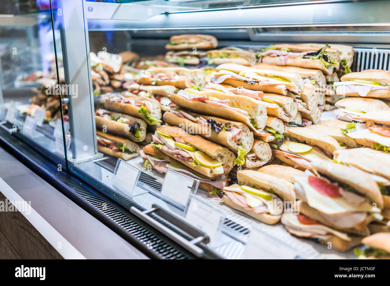 Display of many large sandwiches behind glass window of store stuffed with ham, cheese and cucumbers Stock Photo