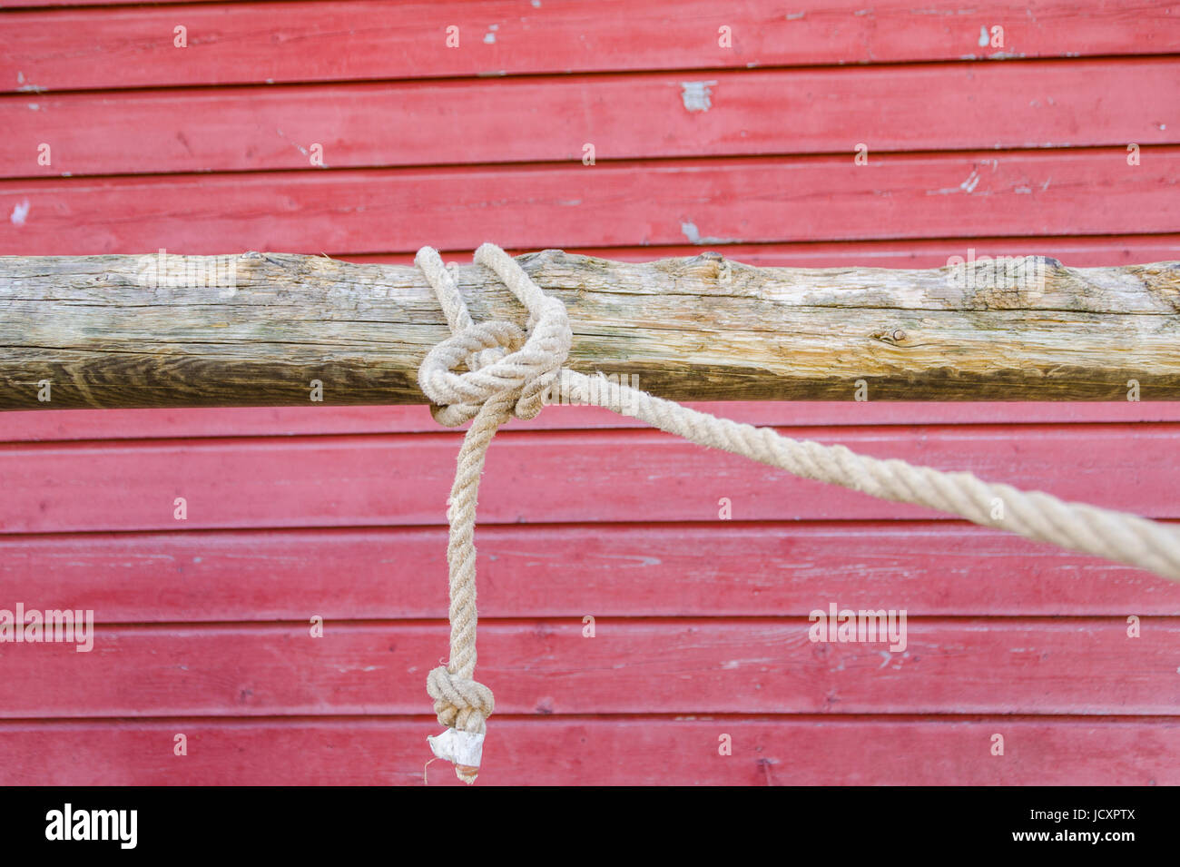 Rope tied in a knot on a hitching post in front of a rustic red barn wall Stock Photo