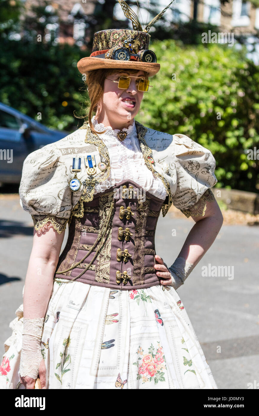 Steampunk fashion. Woman, 20s, standing with hand on hip wearing dress with front lace-up corset, and hat with goggles and feather. Outdoors, bright sunshine. Stock Photo