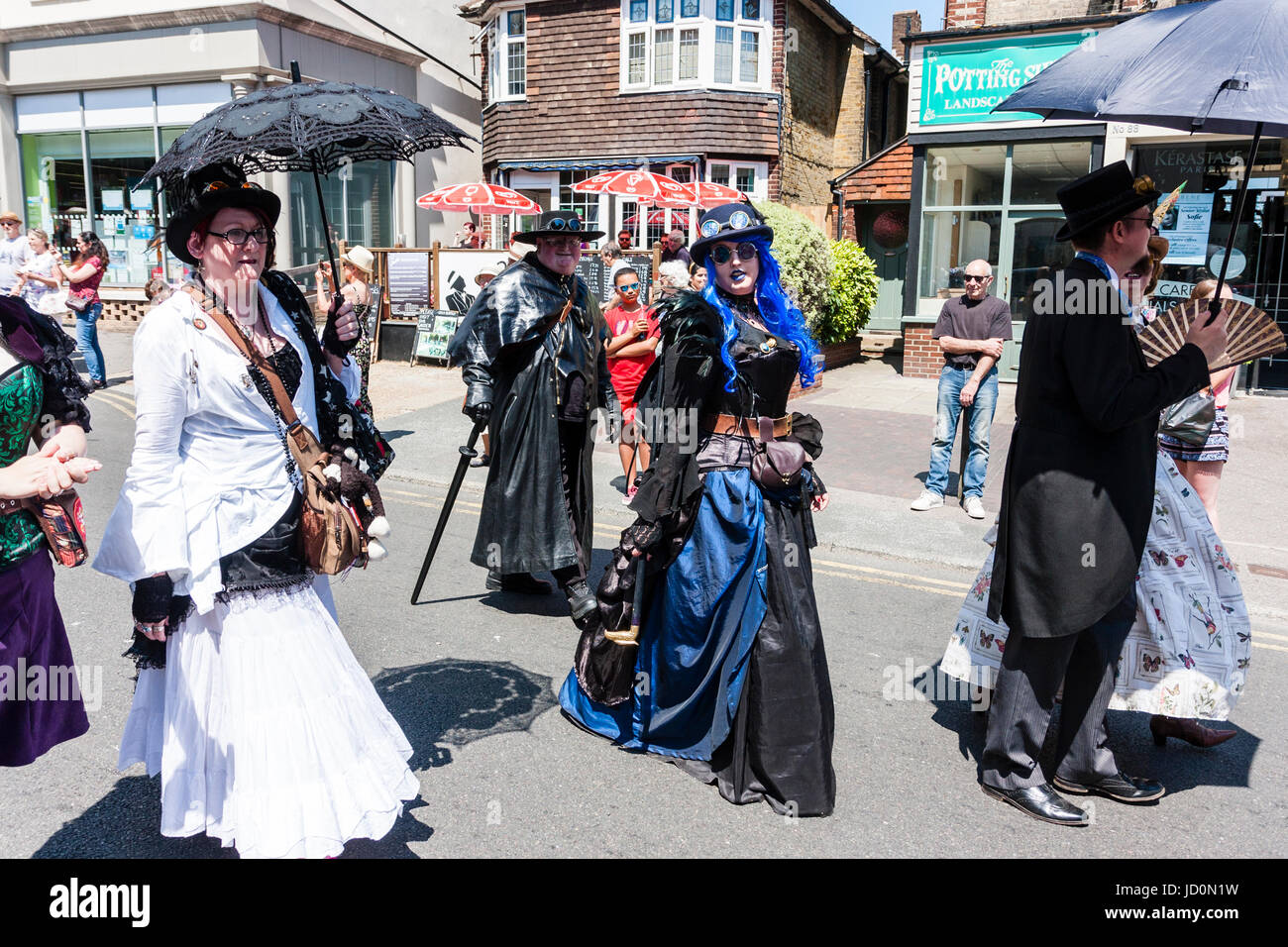 Steampunks marching along street as part of a parade. One, young woman in black and blue gothic type dress, turns to look at viewer as they walk past. Others holding black umbrellas as sunshades. Stock Photo