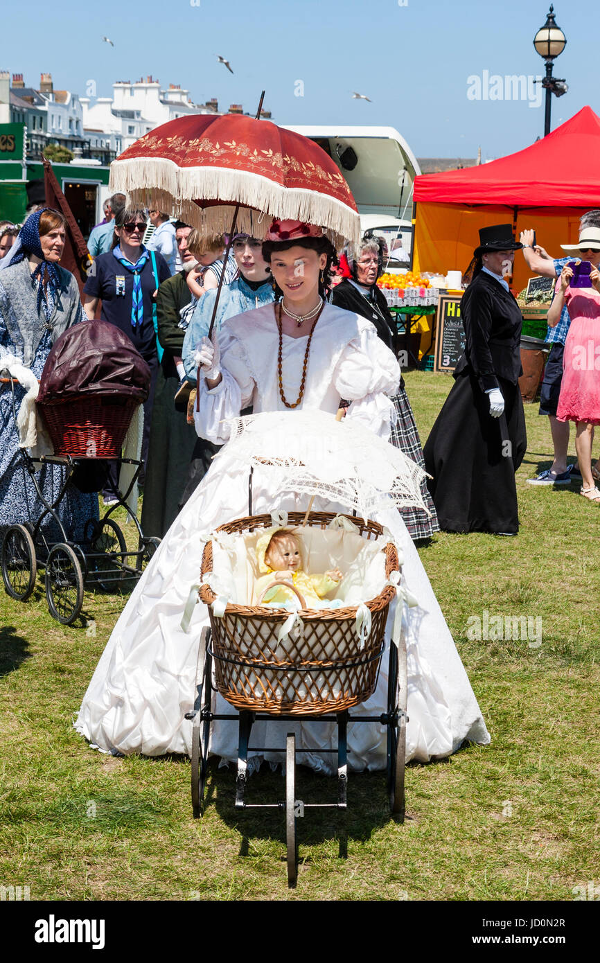Beautiful young woman holding a parasol and pushing a pram with baby basket on and doll laying inside. Dressed in Victoria long dress, part of the Dickens week at Broadstairs, England. Stock Photo