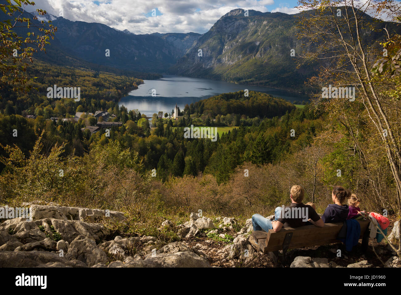 Family with small children resting on a bench admiring the view over a landscape with Lake Bohinj and mountains during a hike in autumn in Slovenia. Stock Photo