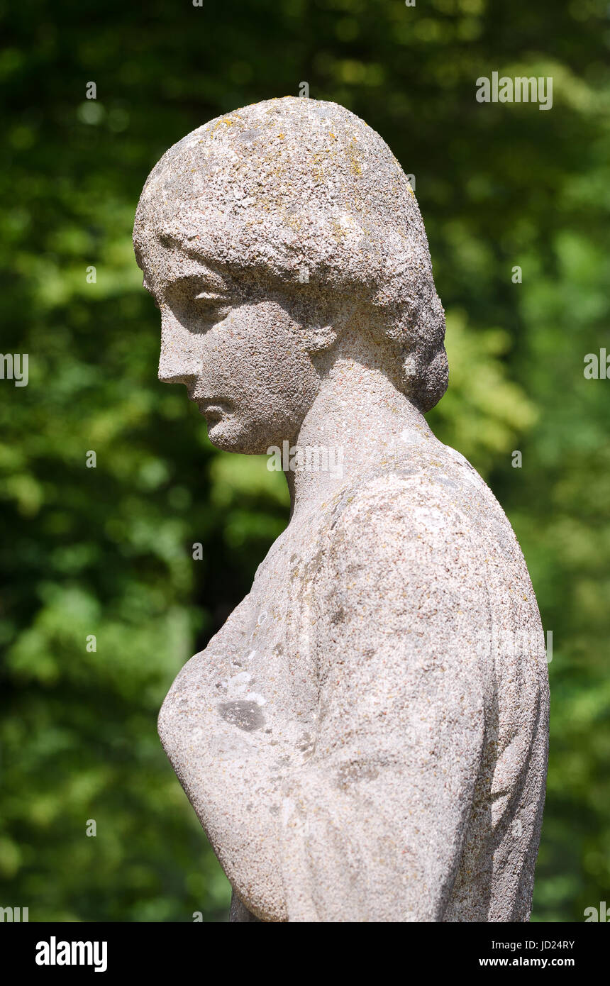 Old stone statue of a woman in sunshine. Her left hand at the bosom, she looks thoughtful, sad, depressed or lonely. Antique weathered sculpture. Stock Photo