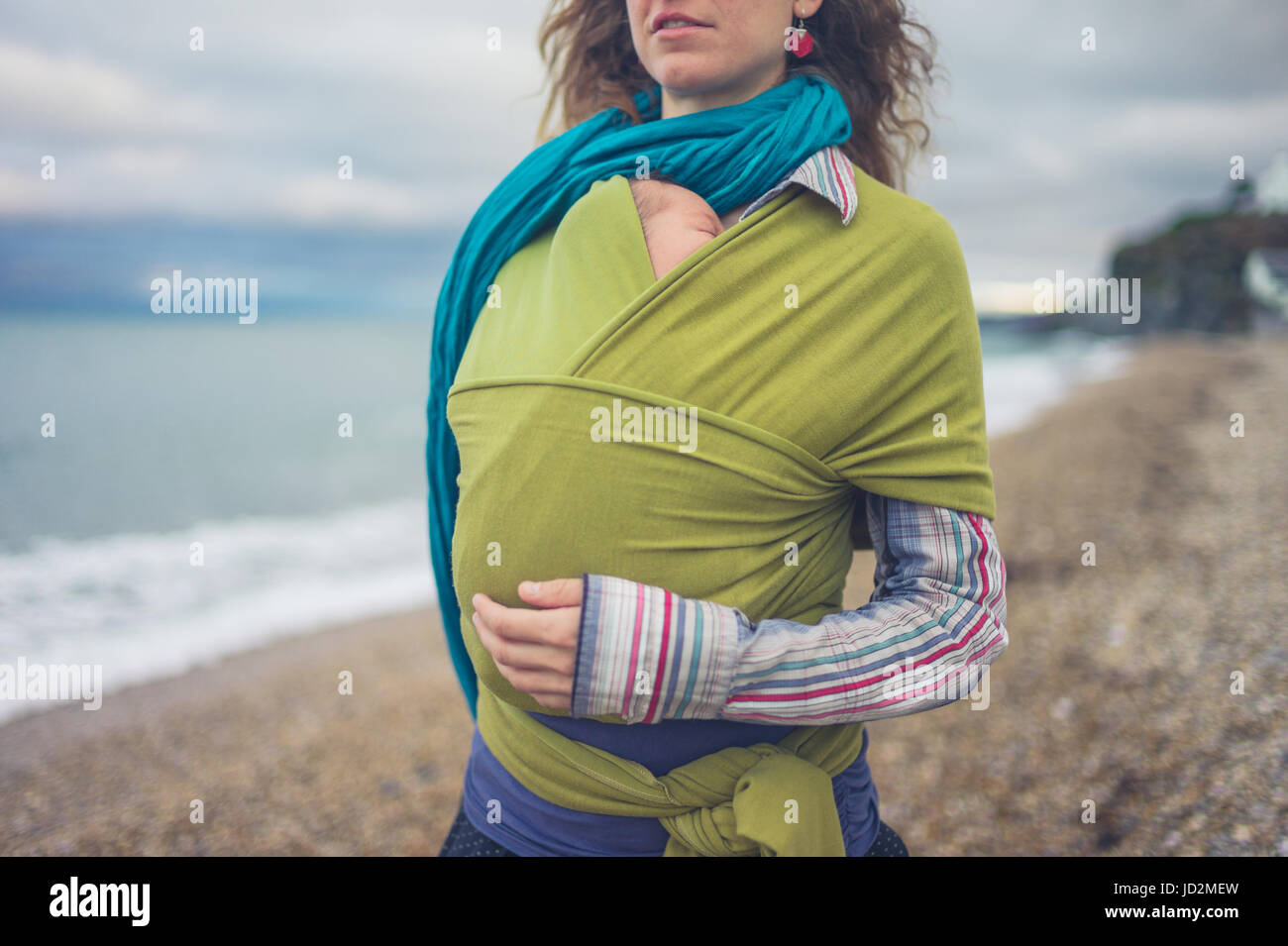A young mother is by the sea with her baby wrappedd in a sling Stock Photo
