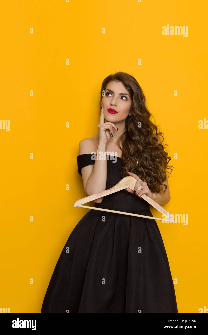 Beautiful young woman in elegant black cocktail dress is holding wooden coat hanger, thinking and looking up. Three quarter length studio shot on yell Stock Photo