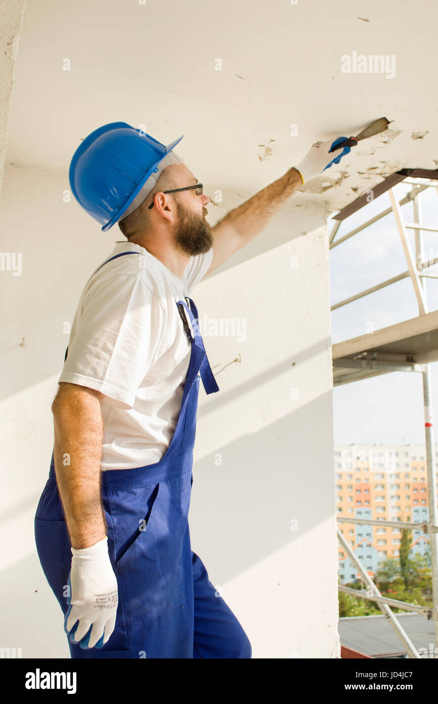 Construction worker in work attire, protective gloves and a helmet on site. Remove the old paint spatula from the ceiling. Stock Photo