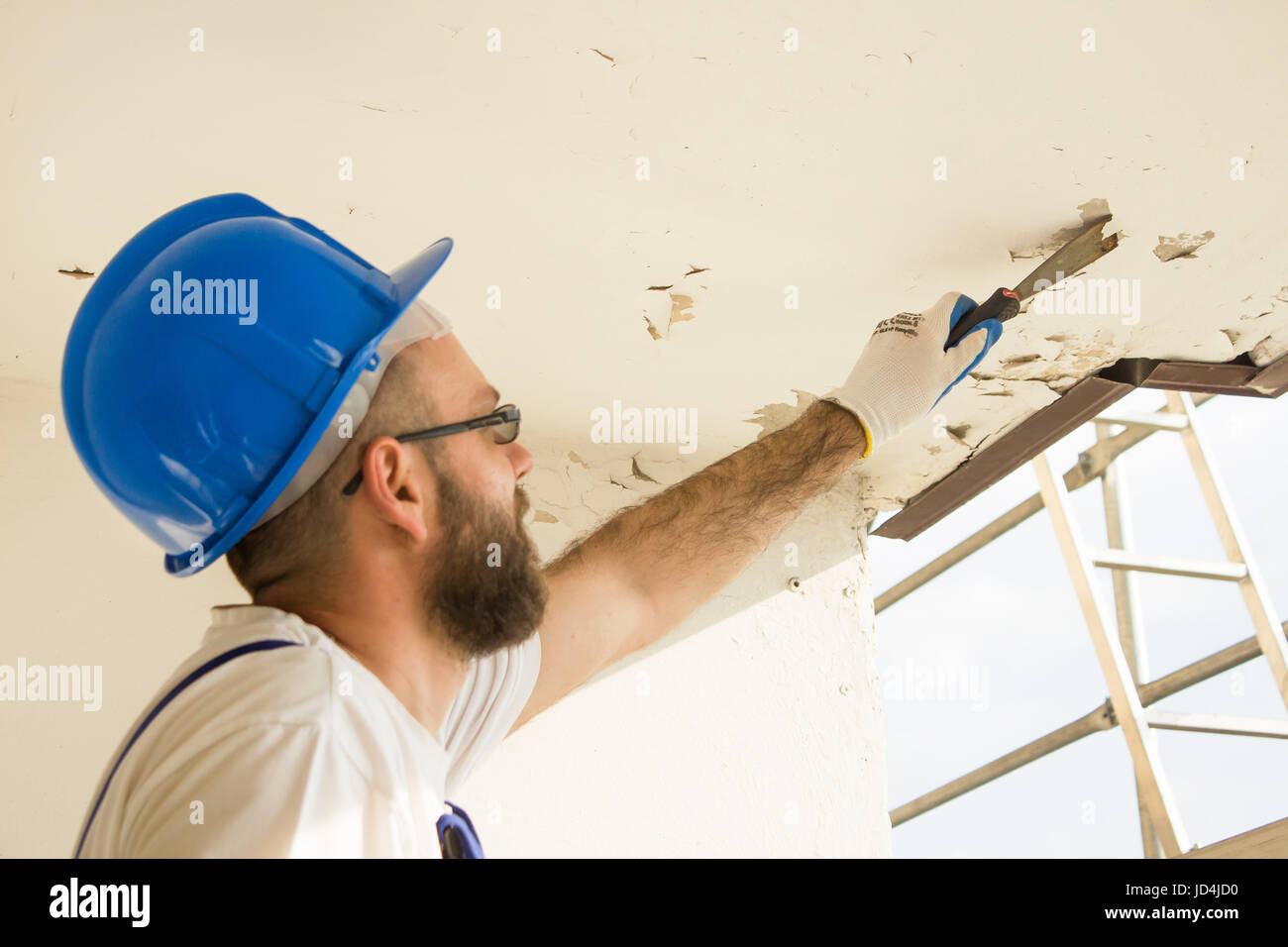 Construction worker in work attire, protective gloves and a helmet on site. Remove the old paint spatula from the ceiling. Stock Photo