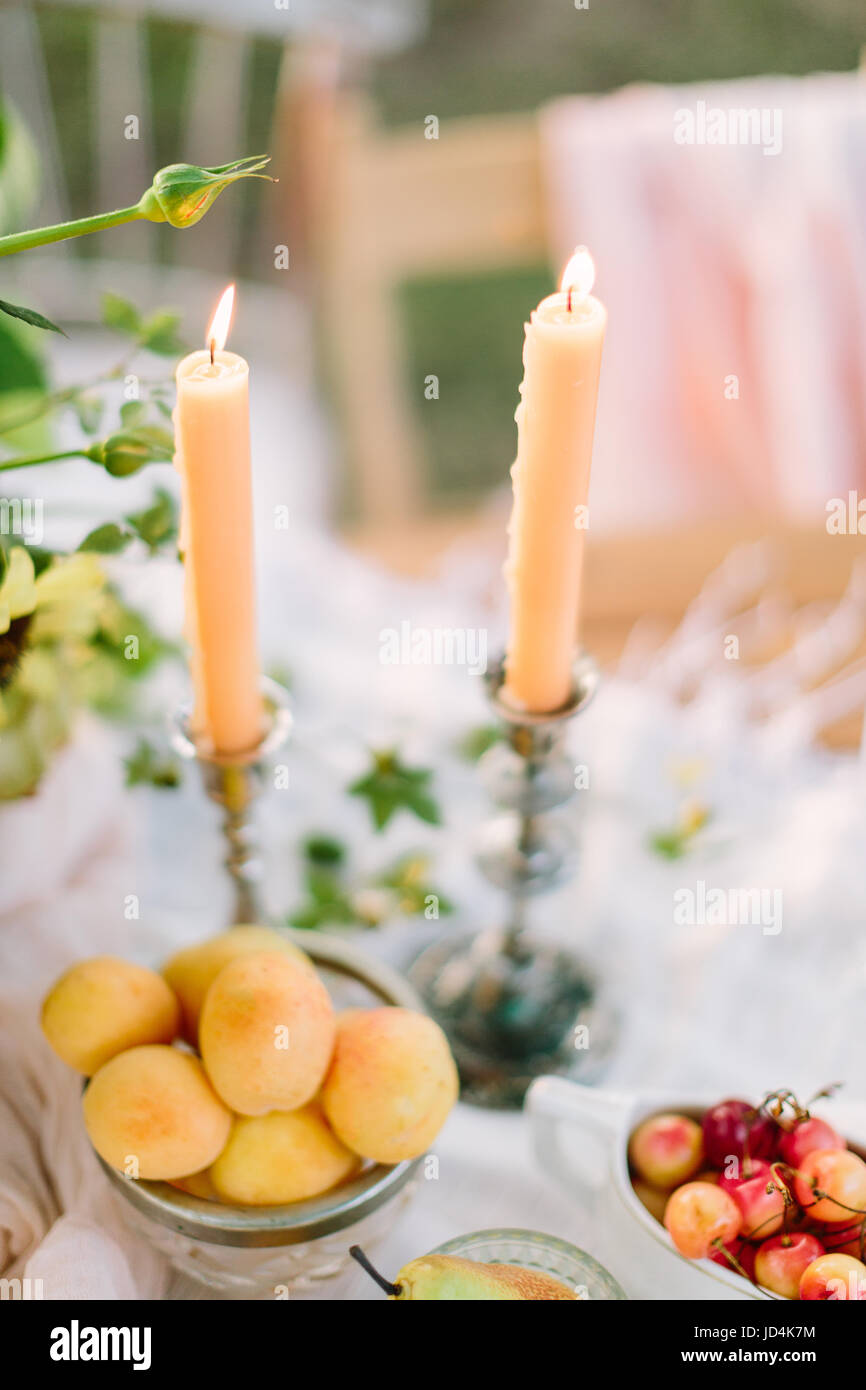 dating, healthy life, romance, wedding, decoration, celebration concept - still-life with two slender candles, apricots in white bowl with gold band and red-yellow wild cherries Stock Photo