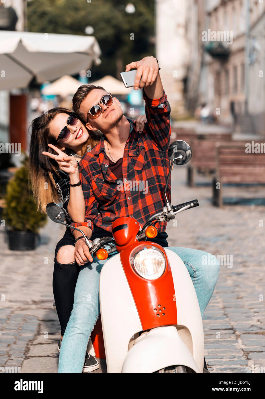 Young attractive couple of teenagers wears sunglasses on retro motorbike makes selfie, sunny street, urban hipster concept Stock Photo