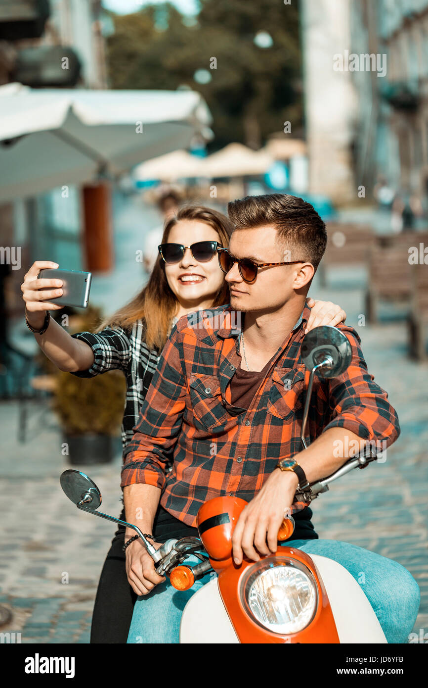 Young attractive couple of teenagers wears sunglasses on retro motorbike makes selfie, sunny street, urban hipster concept Stock Photo