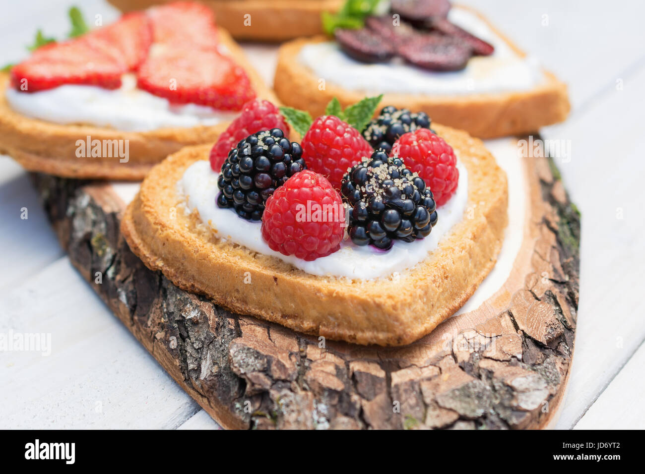 Heart shaped biscuits spread with quark, strawberries, blackberries, raspberries, cherries and a twig of mint presented on a tree disk Stock Photo