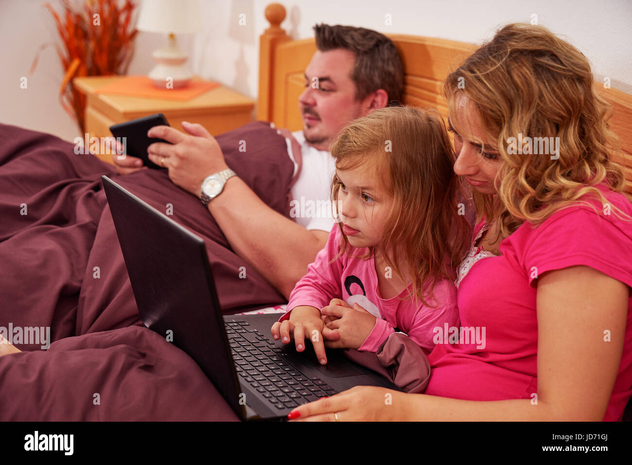 A family (mother, father, girl) lying in bed and using gadgets, wenn girl looking something astounding in notebook Stock Photo