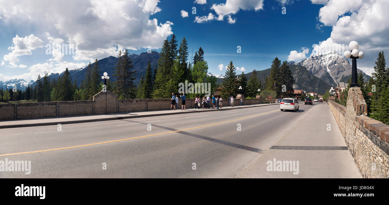 Sunny panoramic street scenery of people on Banff Avenue bridge in the Town of Banff in Alberta Rockies with Rocky Mountains in the background. Albert Stock Photo