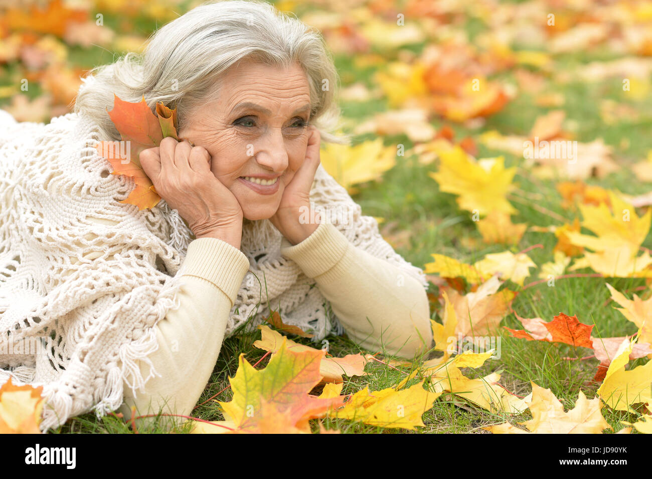 Beautiful senior woman lying on green grass with colorful autumn leaves Stock Photo