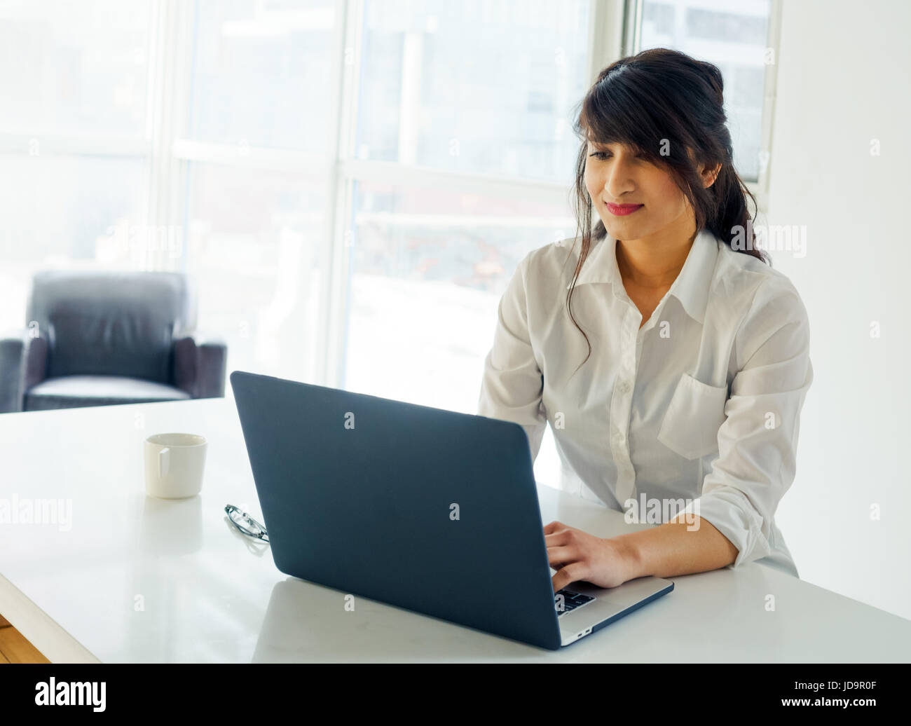 Young woman using laptop at desk, day, with window behind. young adult arabic pretty 20 years old Stock Photo