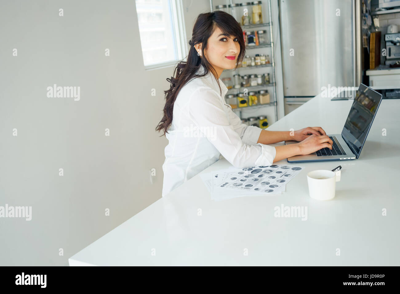 Young woman looking at camera, using laptop in kitchen at home. young adult arabic pretty 20 years old Stock Photo
