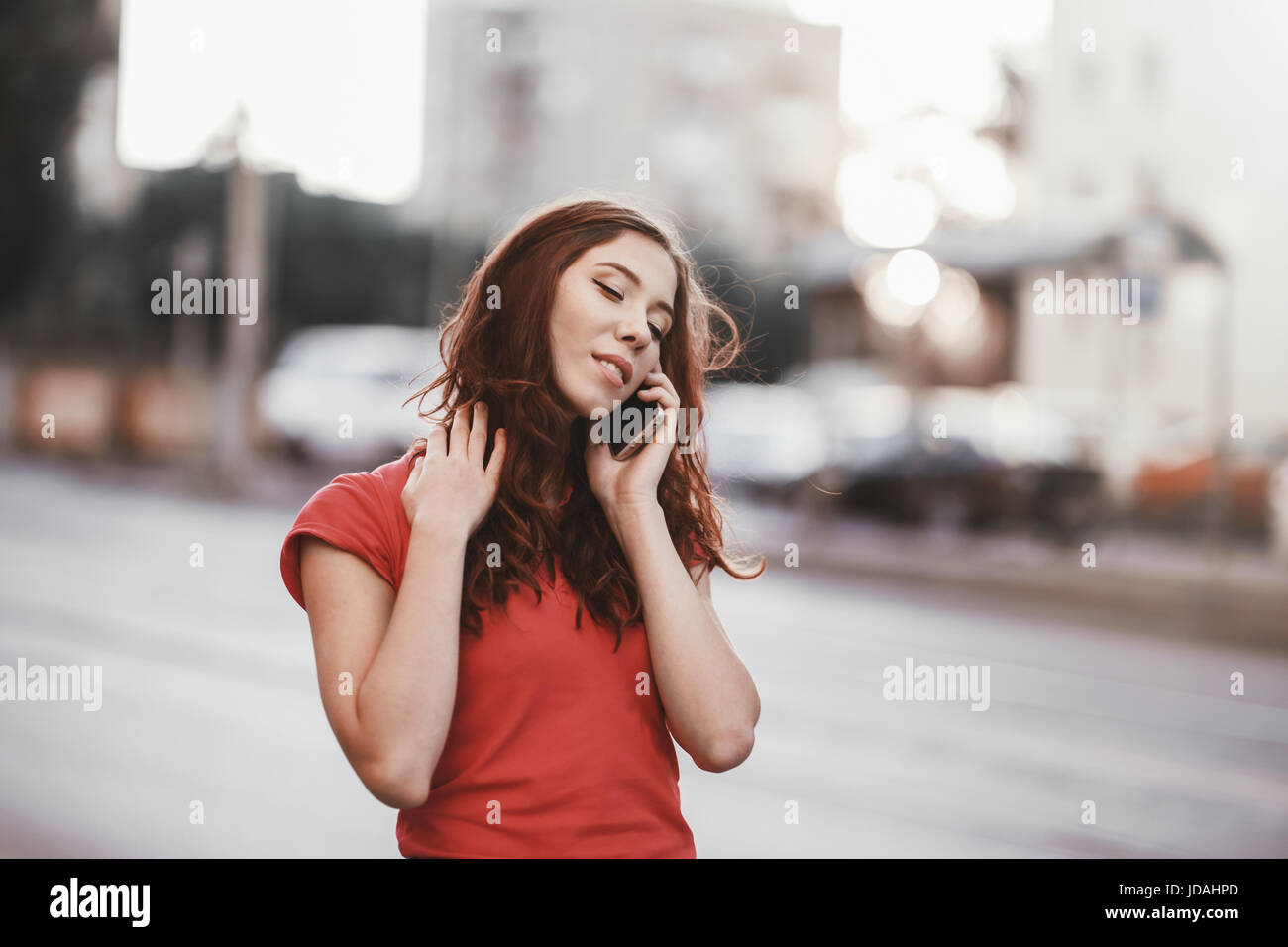 Pretty girl in casual dress is talking on a mobile phone while walking at the city street on a sunset time. Stock Photo