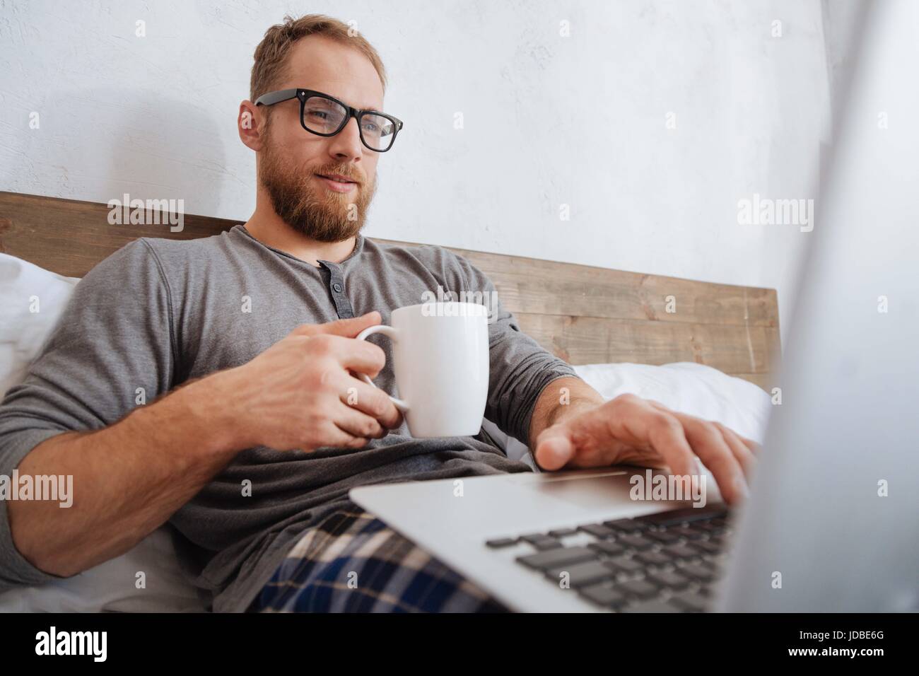 Cheerful man drinking coffee and working with laptop in bed Stock Photo