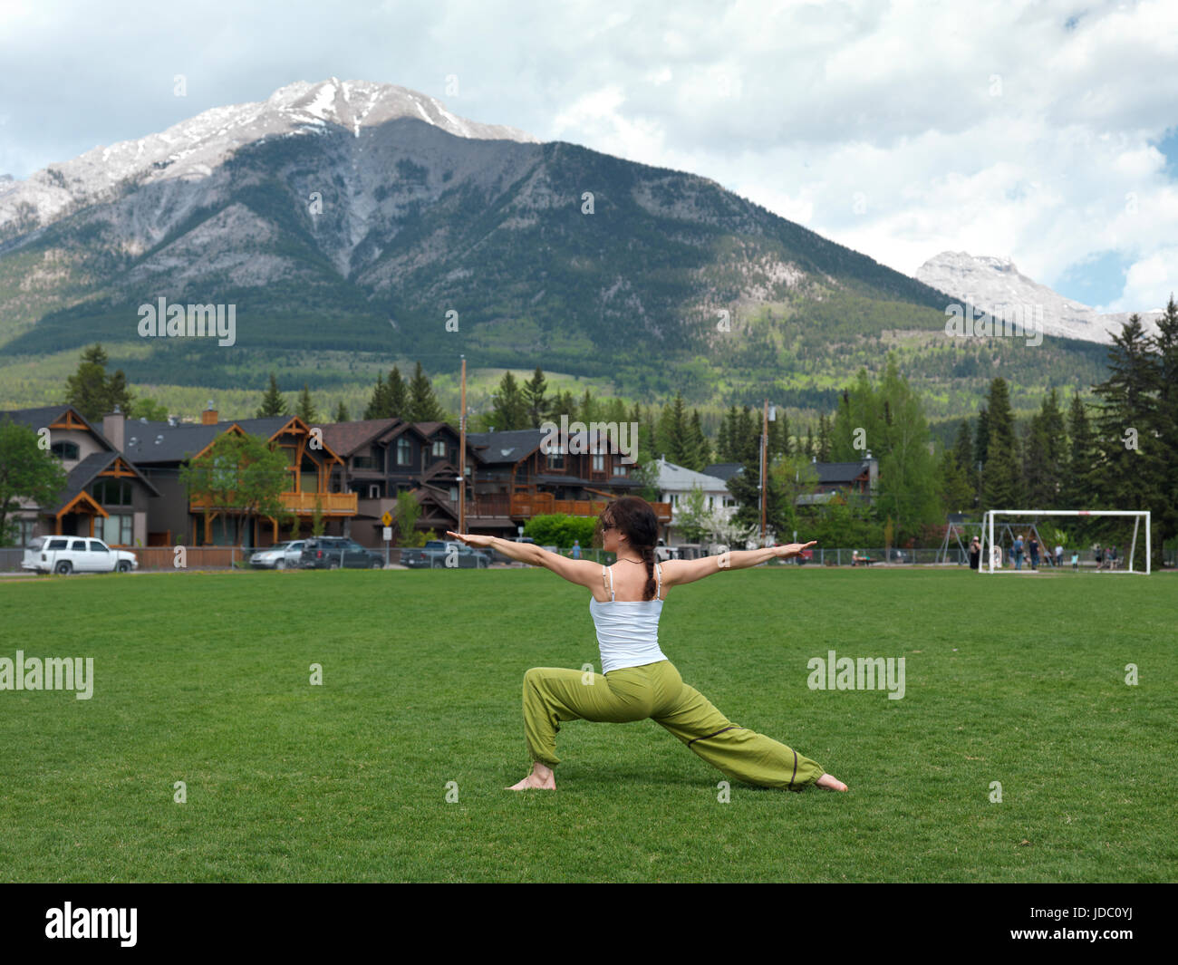 Young woman practicing yoga warrior posture outdoors in Centennial Park in Canmore, town in Alberta's Rockies with Rocky mountains in the background.  Stock Photo