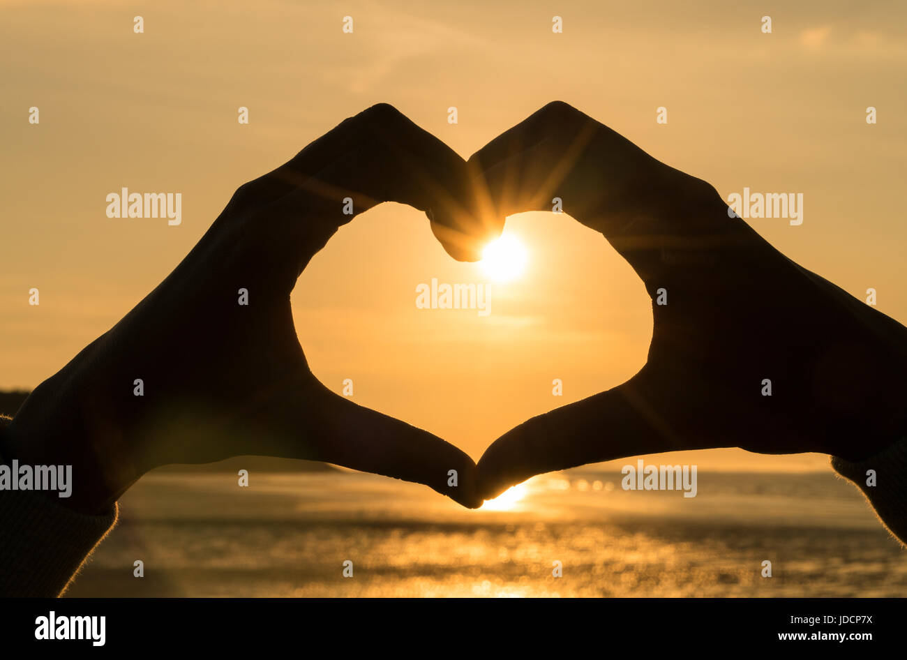 Hand heart frame shape silhouette made against the sun & sky of a sunrise or sunset on a deserted empty beach Stock Photo