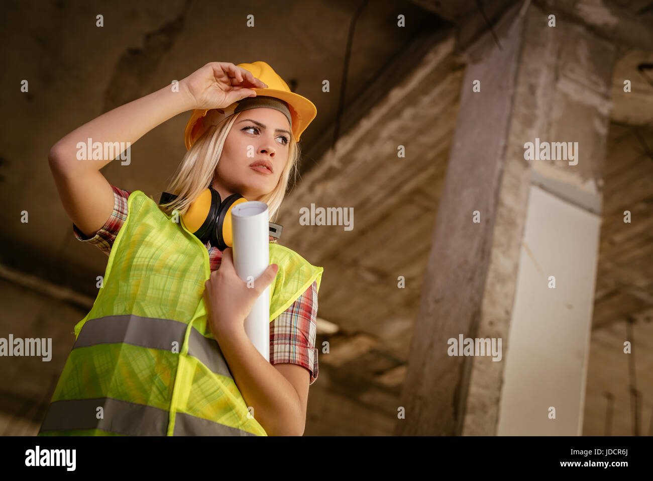 Beautiful young female construction architects at a construction site. She is holding plans and looking away. Stock Photo