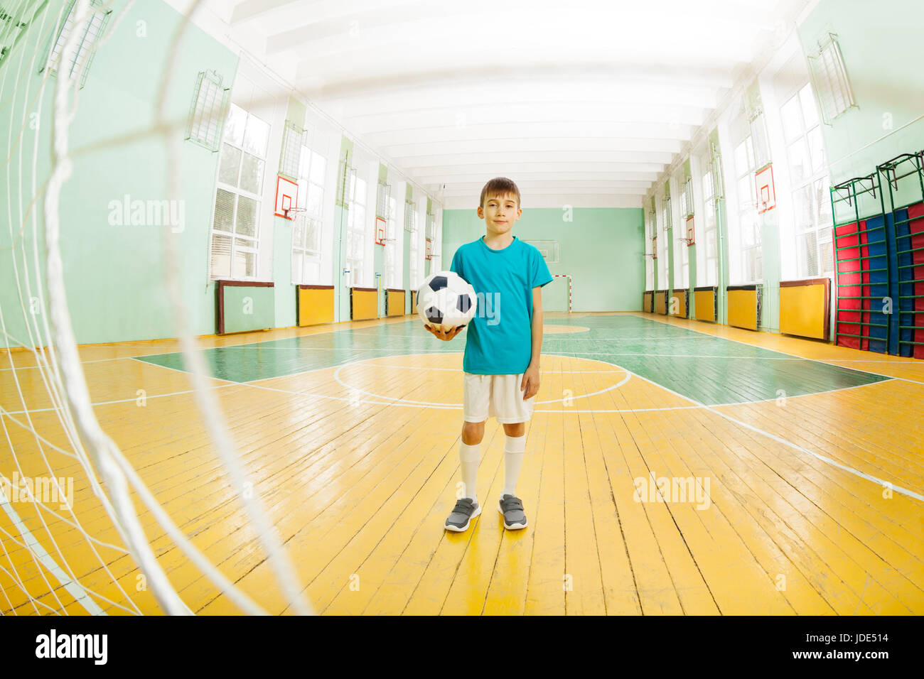 Full-length portrait of 12 years old boy in football uniform, standing in school gymnasium, holding soccer ball, shot through the gate net Stock Photo