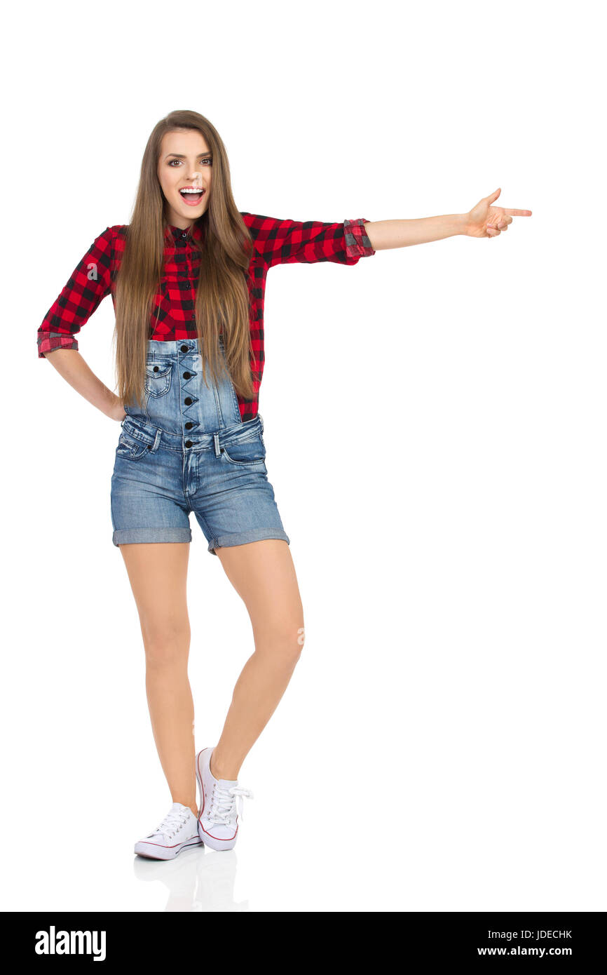Shouting young woman in red lumberjack shirt, jeans dungarees shorts and  white sneakers standing with hand on hip pointing and looking at camera.  Full Stock Photo - Alamy