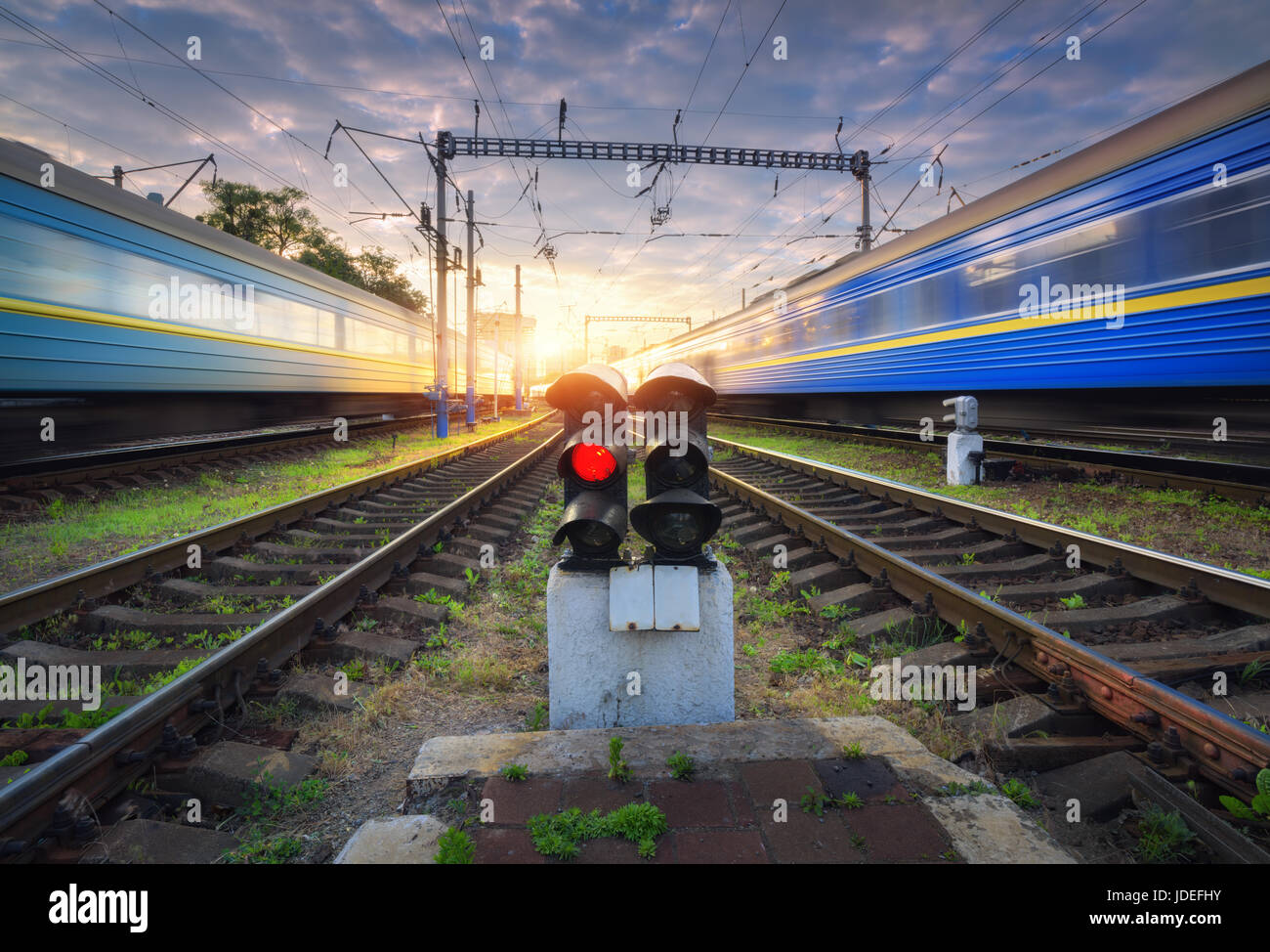 High speed passenger trains in motion on railroad track at sunset. Railway station with blurred modern commuter train, railway traffic light, blue clo Stock Photo