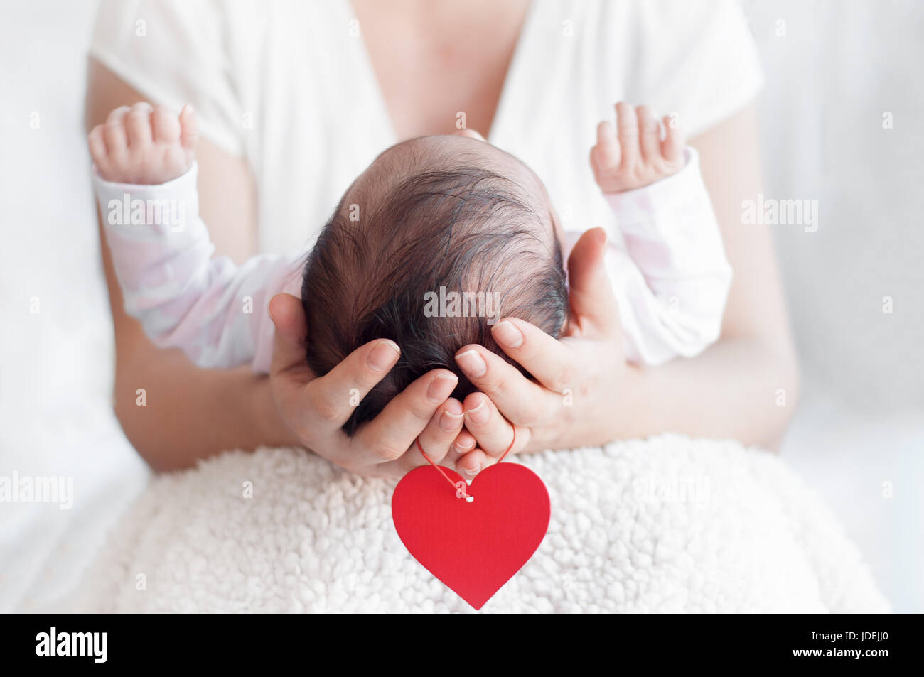Mother holding head of her newborn baby in hands. Happy family concept. Maternal love and parental love.  Red heart hanging from the arms. Stock Photo