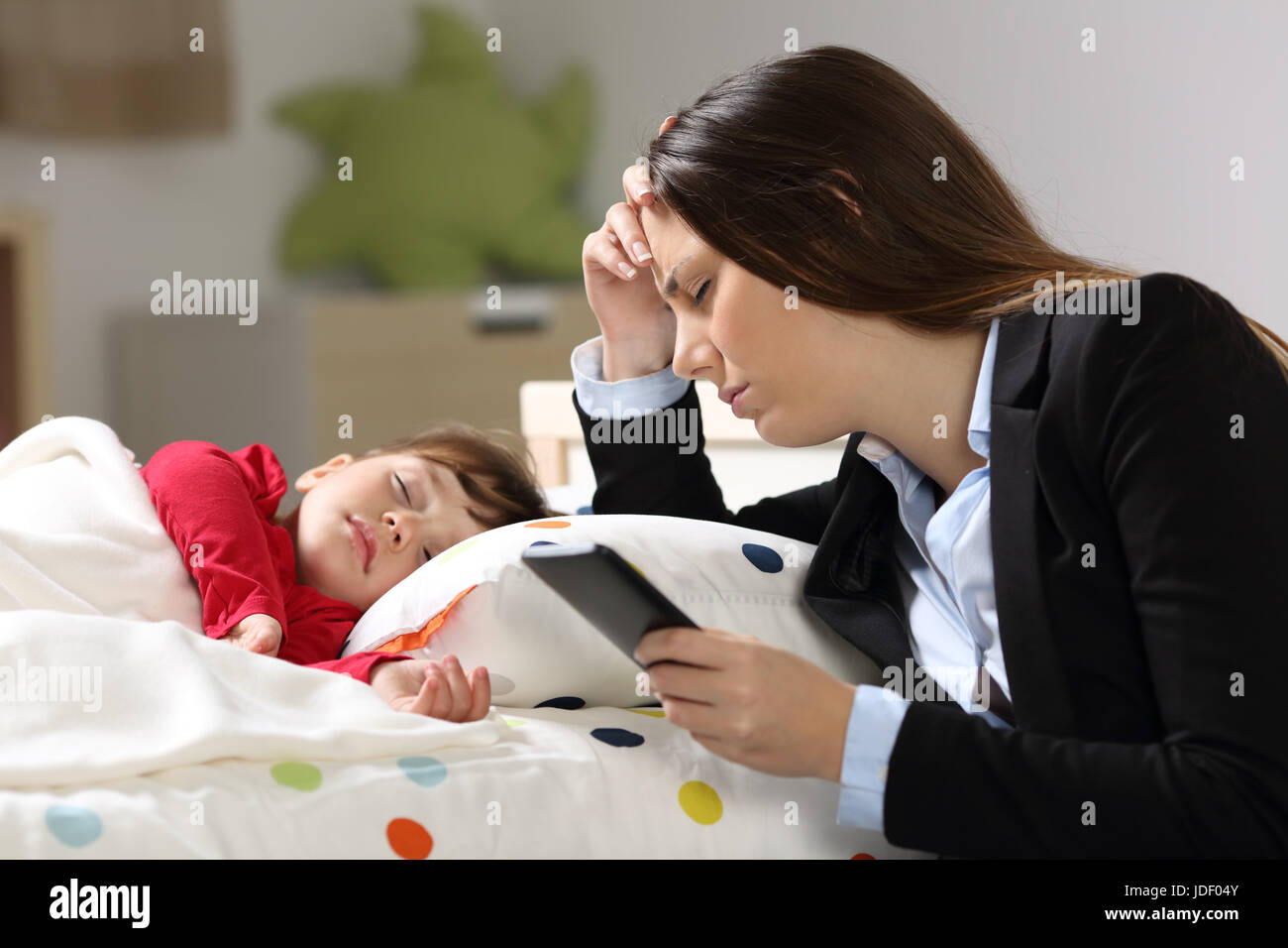 Tired worker mother wearing suit after work while her daughter is sleeping on a bed in a house interior Stock Photo