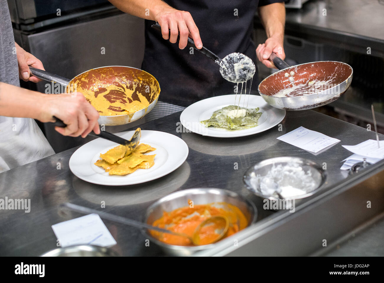 close-up of two unrecognizable chefs in aprons standing next to table in restaurant kitchen and serving ravioli pasta in plates from frying pans Stock Photo