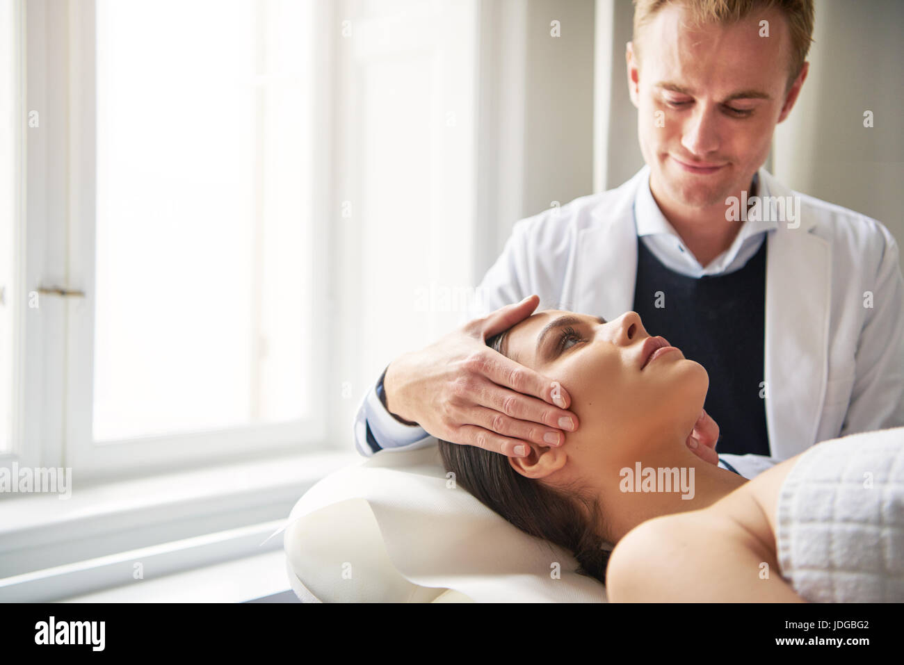 Woman lying on procedure in salon while young cosmetologist man holding her head. Stock Photo